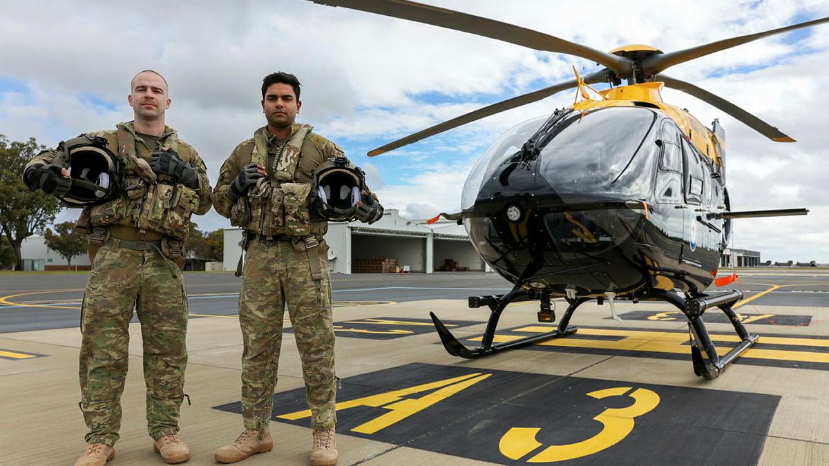 Two military men with helmets stand beside a stationary helicopter on the tarmac.