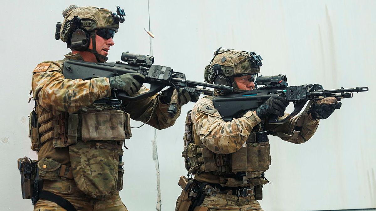 Two men in uniform move alongside a wall with rifles raised, a bullet casing in the air next to one of them.