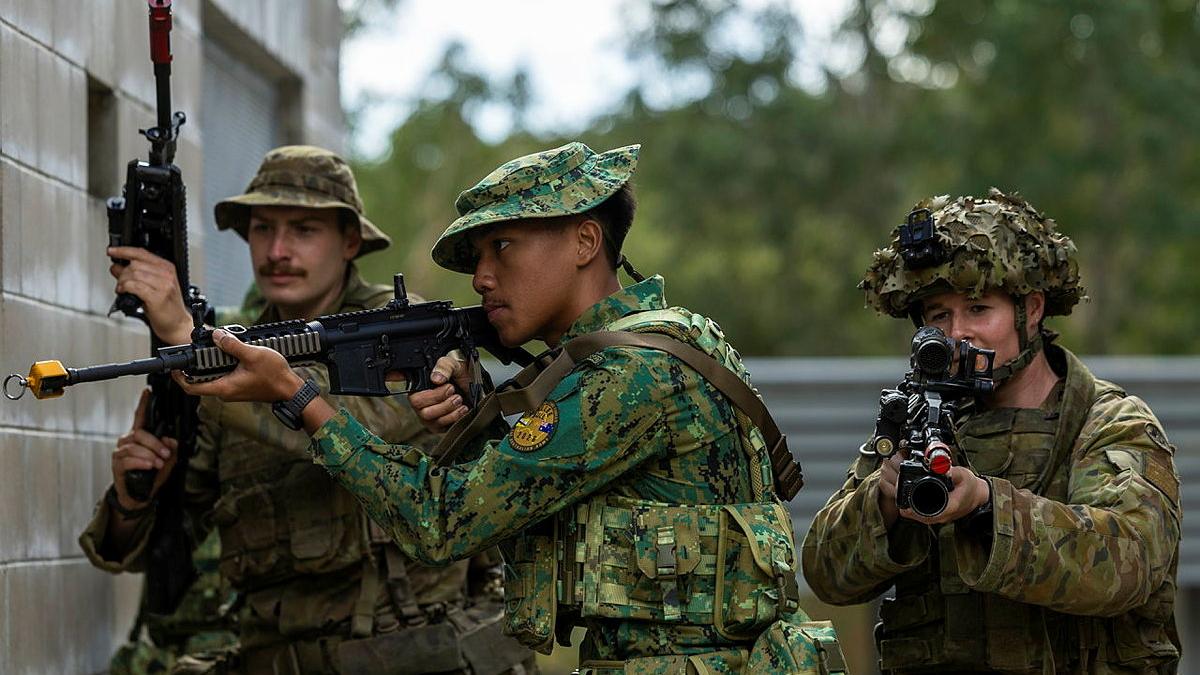 Three people in camouflage uniform hold machine guns, two of them aiming the guns, as they walk next to a cinderblock building.