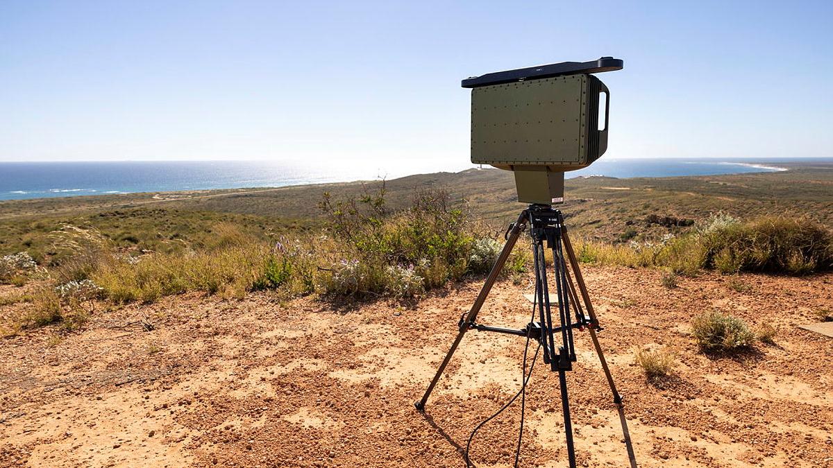 A rectangular device on a tripod standing in red soil, with short scrub nearby and the sea in the distance.