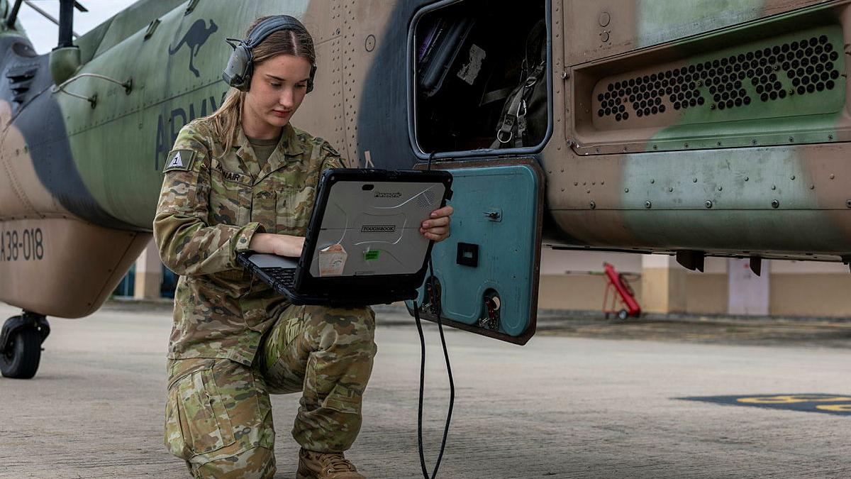 A soldier works with a laptop near a helicopter.
