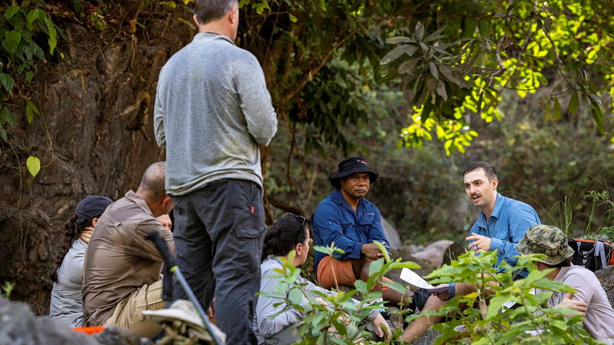 A group of people sit and stand amongst vegetation facing a seated man who is speaking.
