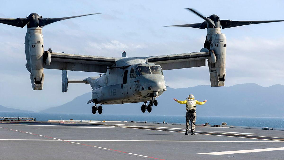A defence member in a bright yellow uniform signal for an aircraft to land on the deck of a ship.