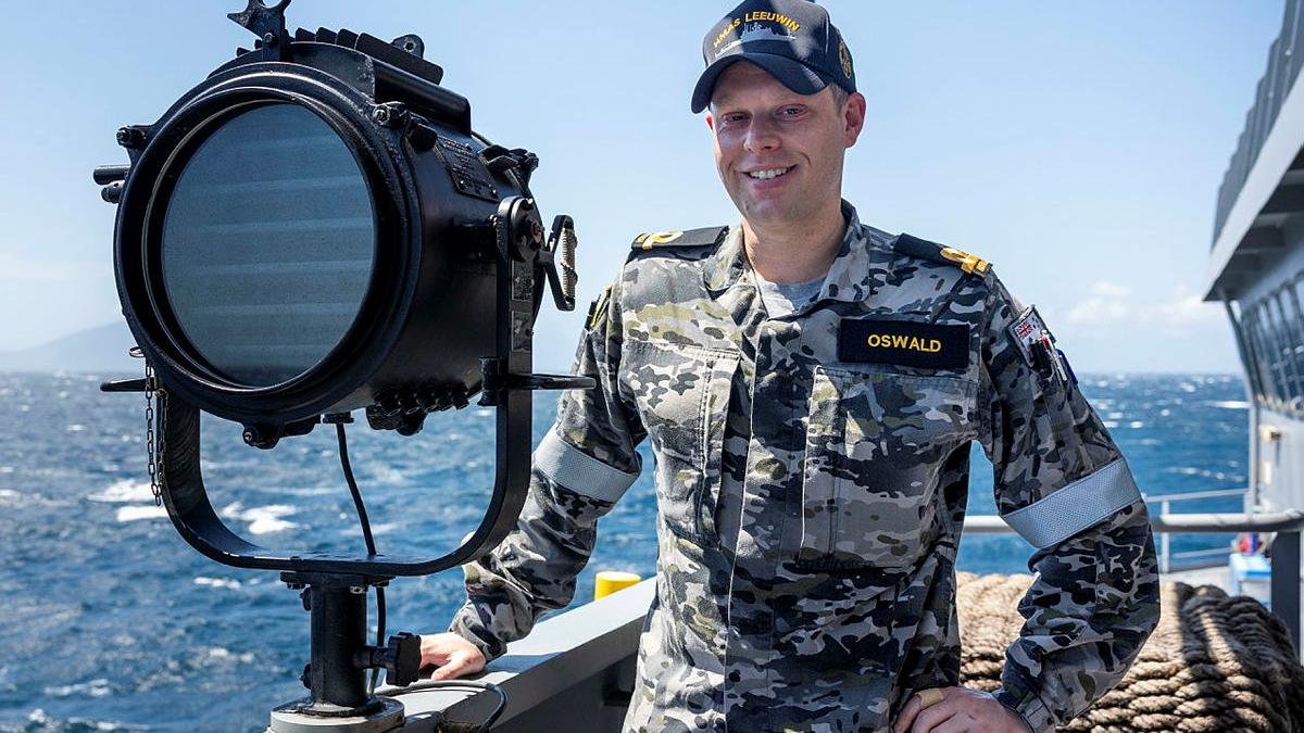A sailor stands on the deck of a ships next to a large lens on a stand.