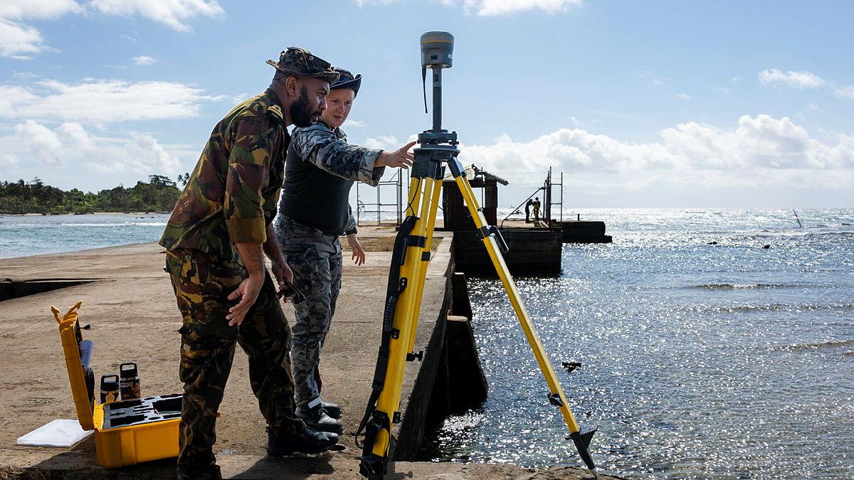 Two people in uniform examine a device on a tripod on a jetty.