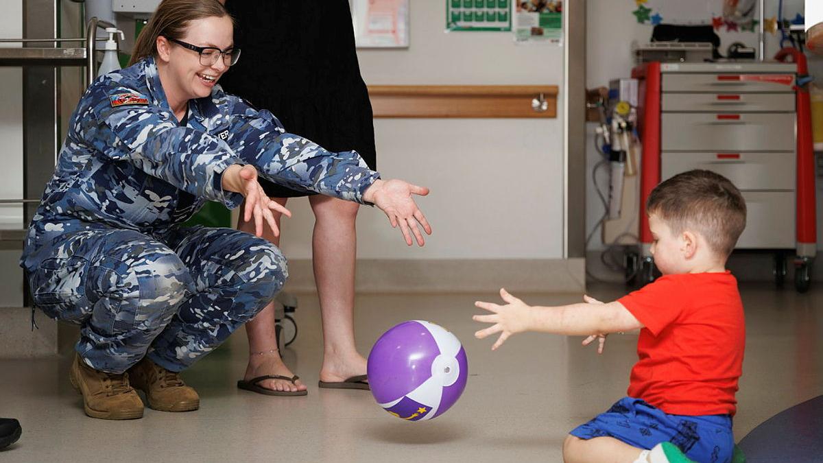 A military woman throws a purple ball to a young child kneeling on the floor.