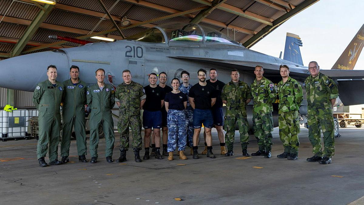 A group of aviators from Sweden, Finland and Australia stand in front of a jet aircraft and smile at the camera.