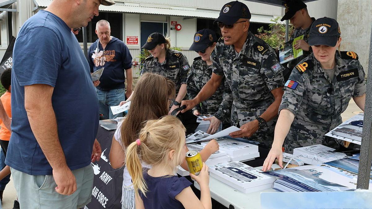 Adults and children talk to Navy personnel while being handed Navy-branded items.