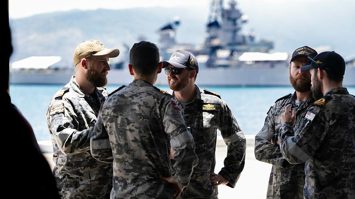 Navy personnel talk in a small group. A ship is seen in the background.