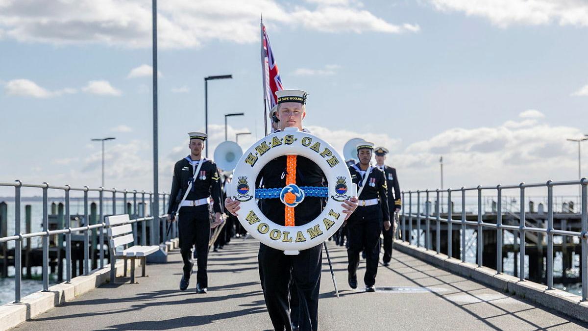A person in Navy uniform carries a lifebuoy along a jetty with a group of uniformed people behind.
