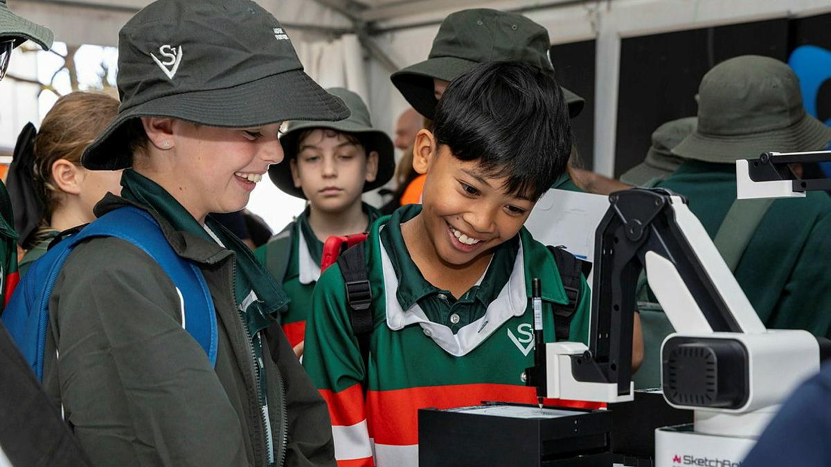 Several young kids inspect a 3D printing machine.