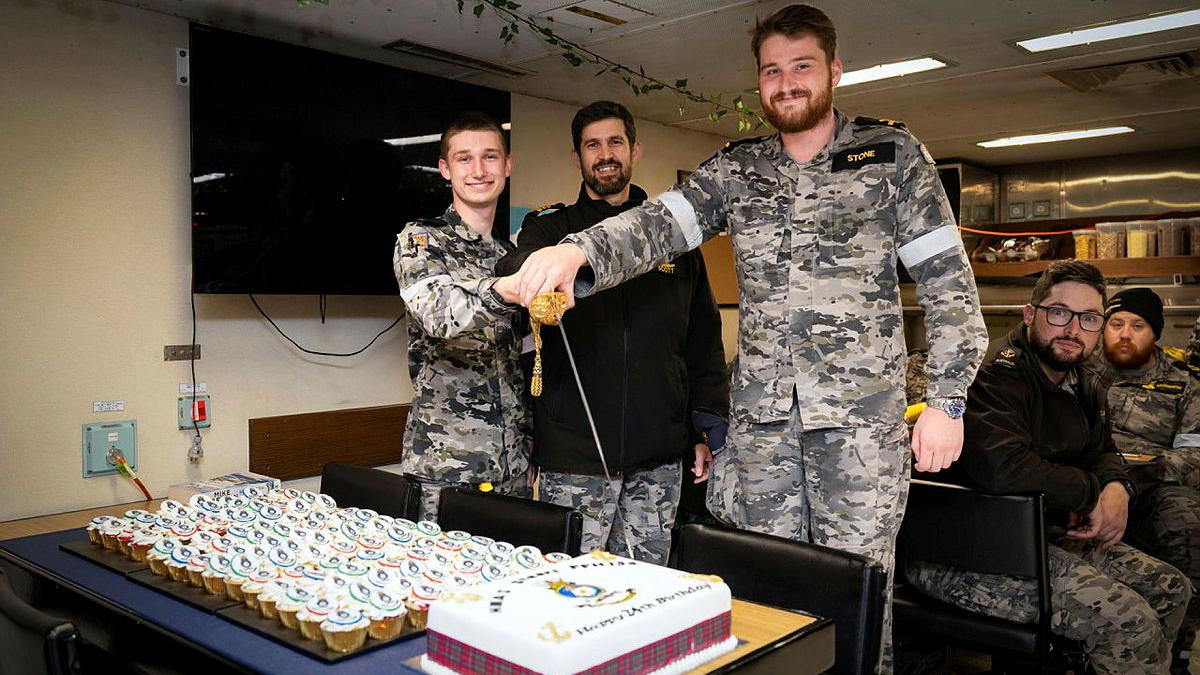 A man in uniform holds a thin sword above a cake while another helps and others watch.