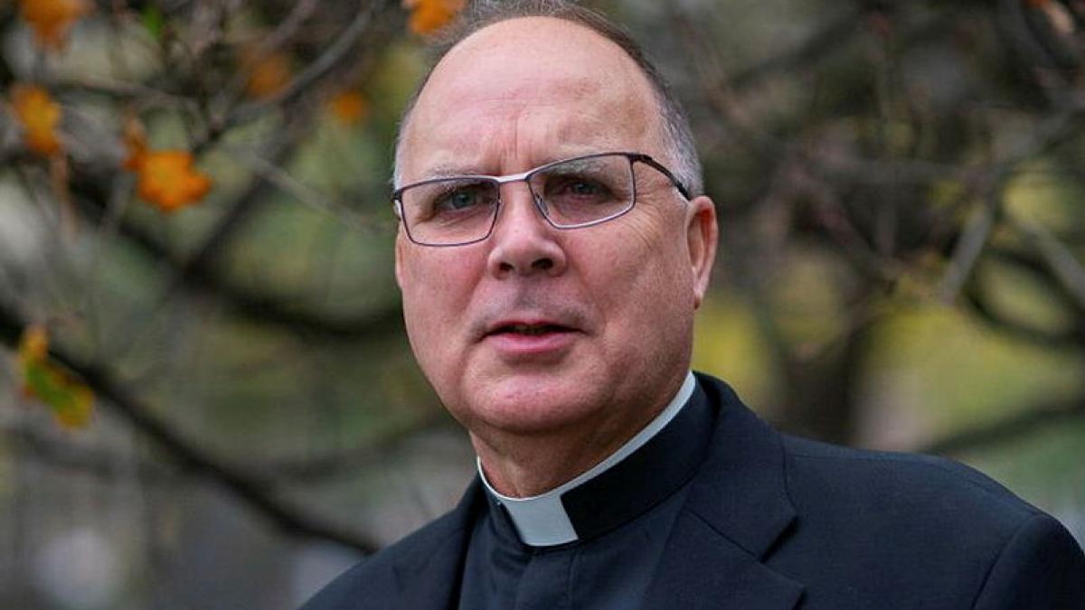 A man in a black suit and clerical collar stands outside.
