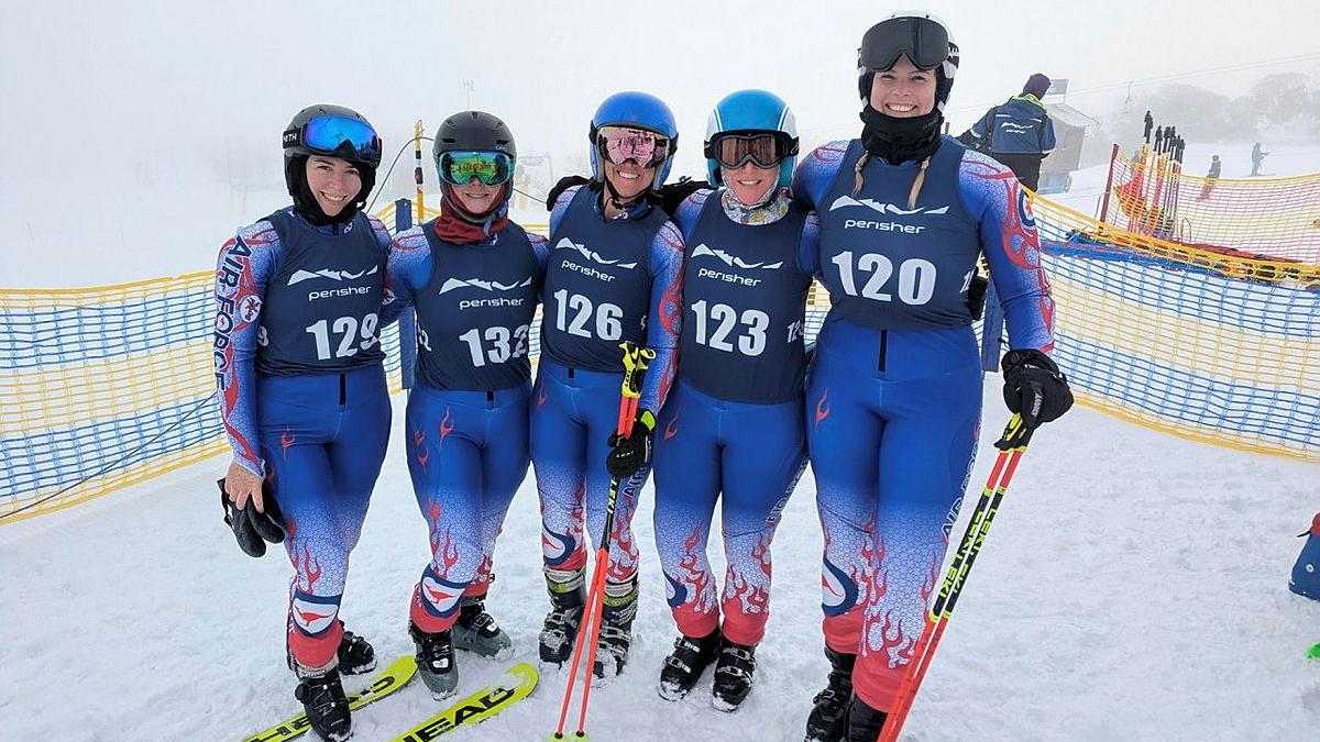 Five women stand together in the snow with snow sport equipment.