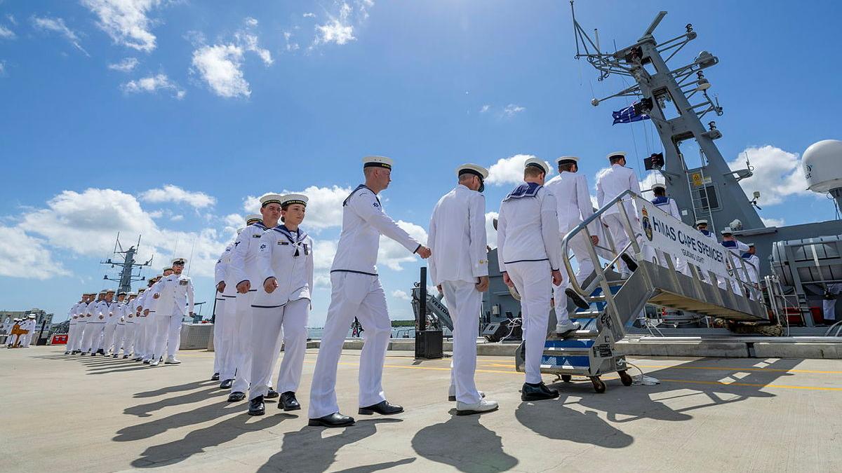 Navy members in white uniforms walk in a line onto a ship on a sunny day.
