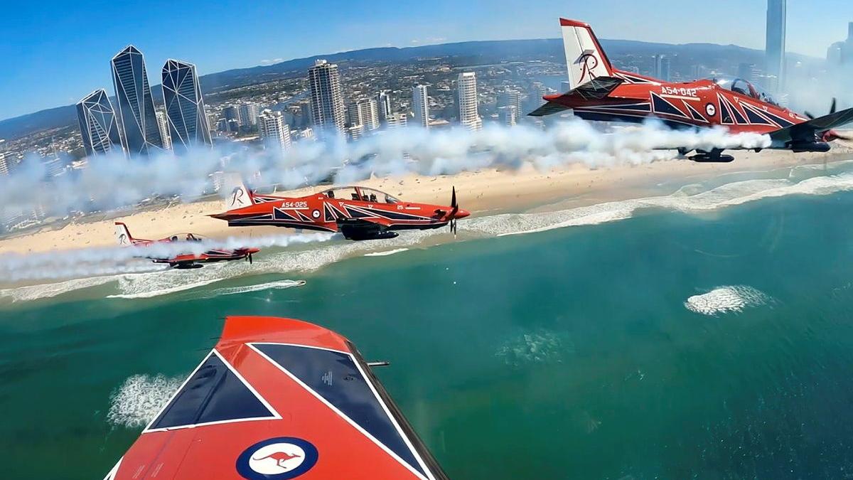 A group of red aircraft fly in formation above a coastal city.
