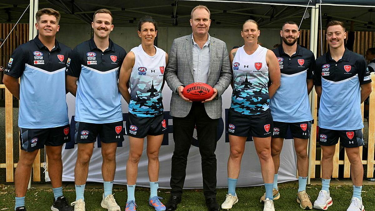Four men in sports uniform, two women in sports uniform and a man in a suit and holding an AFL ball stand in a line.