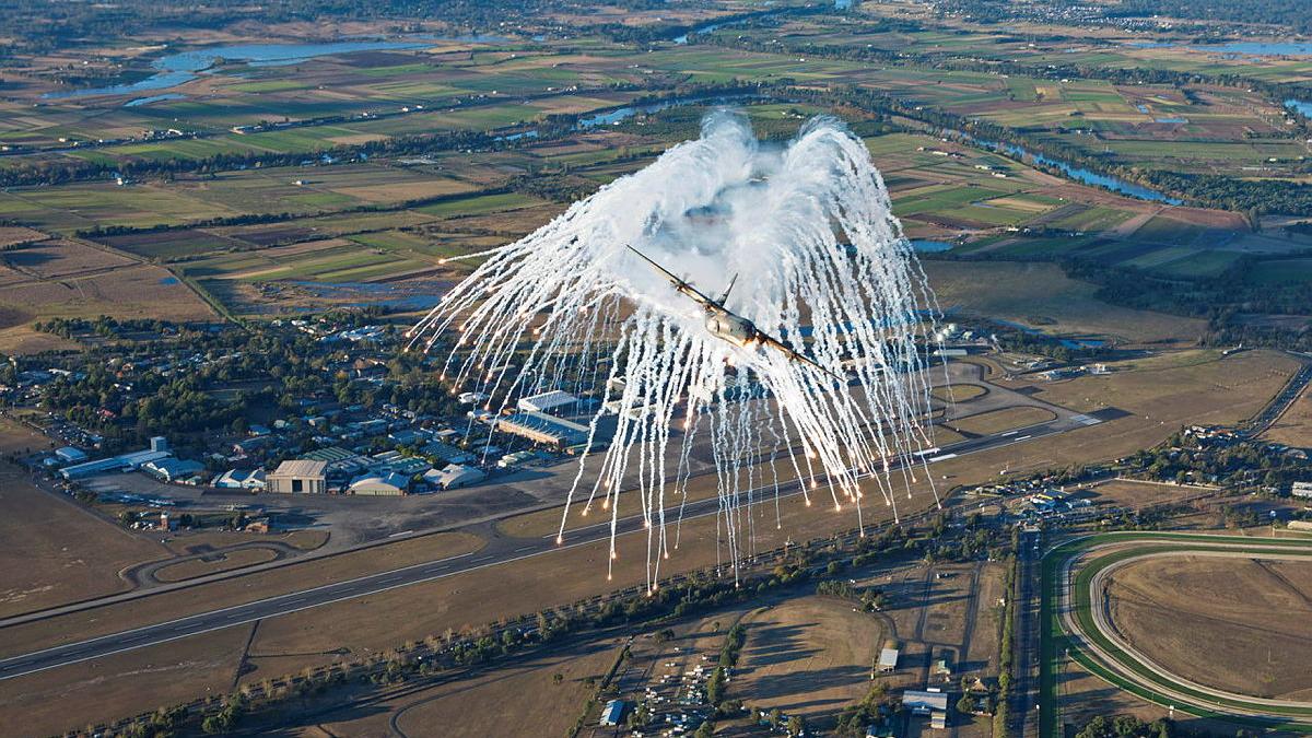 Fireworks explode behind an aircraft in flight over fields and houses.