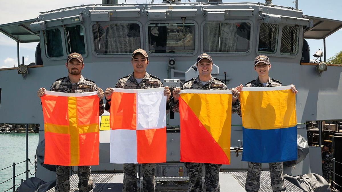 Four Navy personnel from HMAS Diamantina hold up signalling flags on the deck of the ship.
