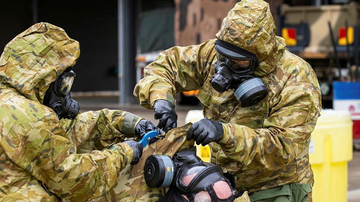 Two people in hazmat suits reach under the hood of a third person in a hazmat suit who is lying down.