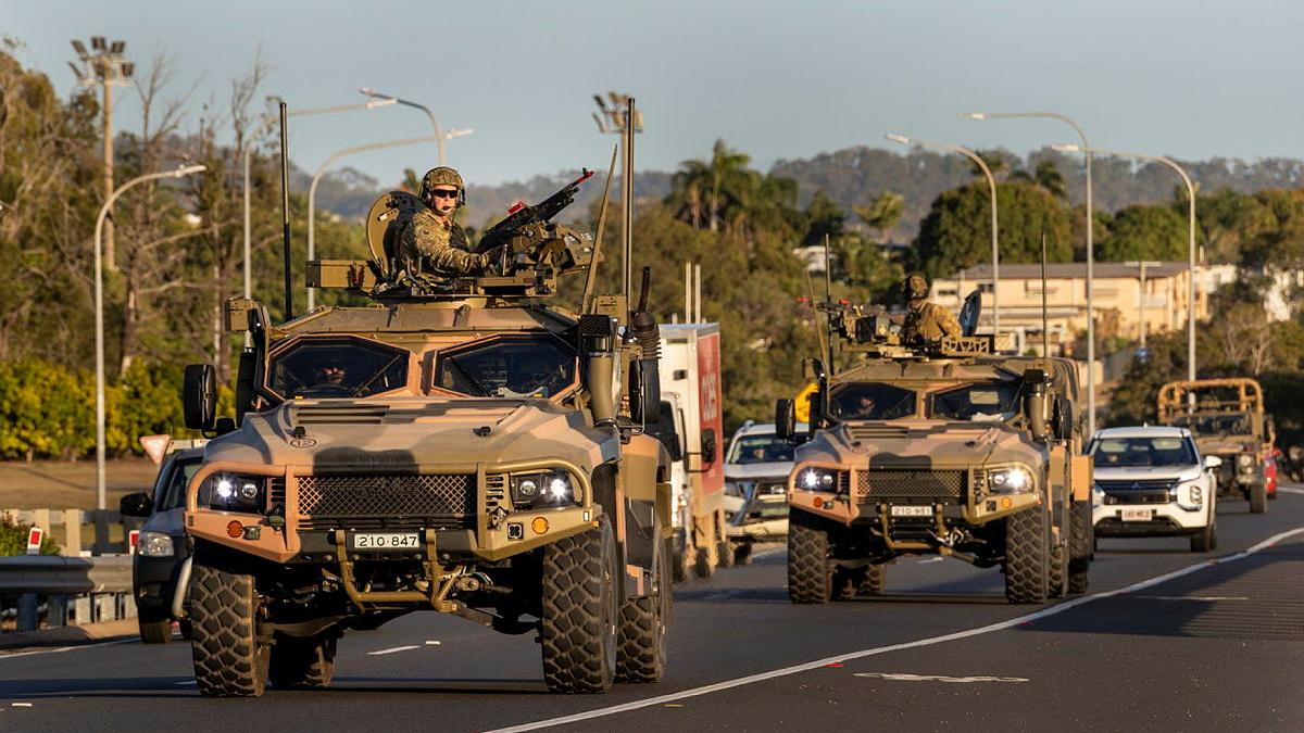 A military man stands in the open hatch of a camo vehicle with an weapon and while the vehicle is driving on the road with other vehicles.