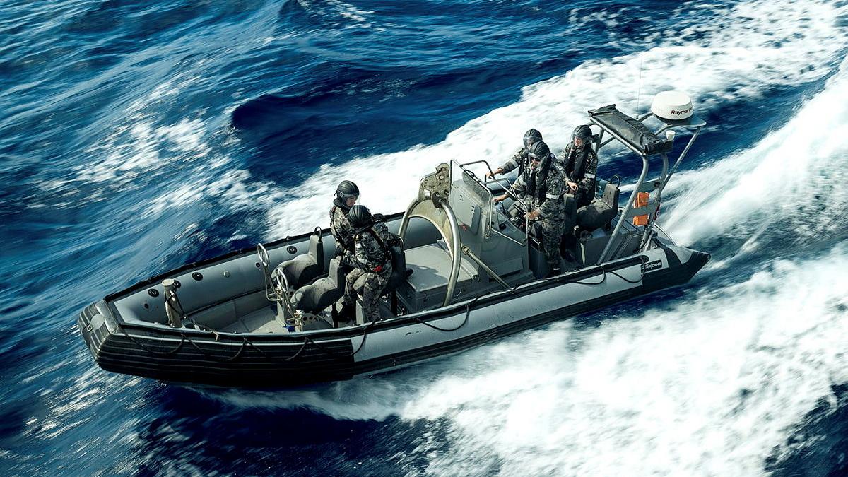 A boat with a small group of people on board in Navy uniforms is surrounded by white foamy water in a deep blue sea.