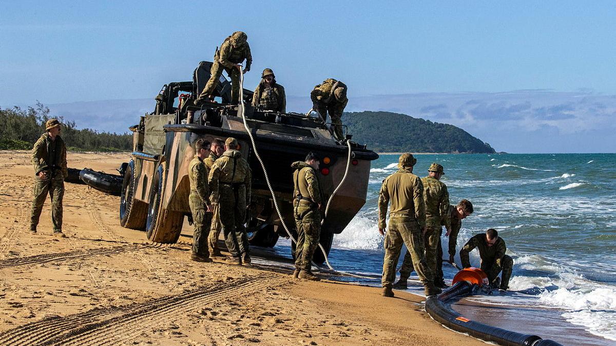 A group of Army members work on and around an Army vehicle with ropes and cables, on the shore of a beach.