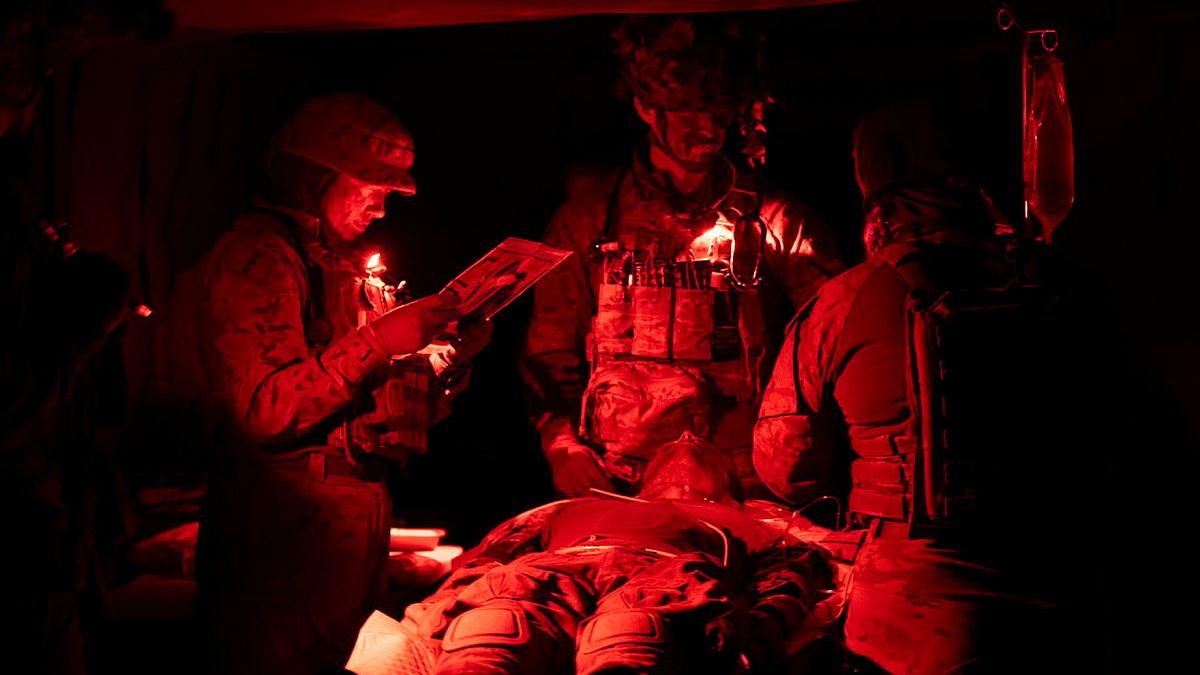 Three people in Army clothing stand under a red light and focus on a person laying down on a table in front of them.