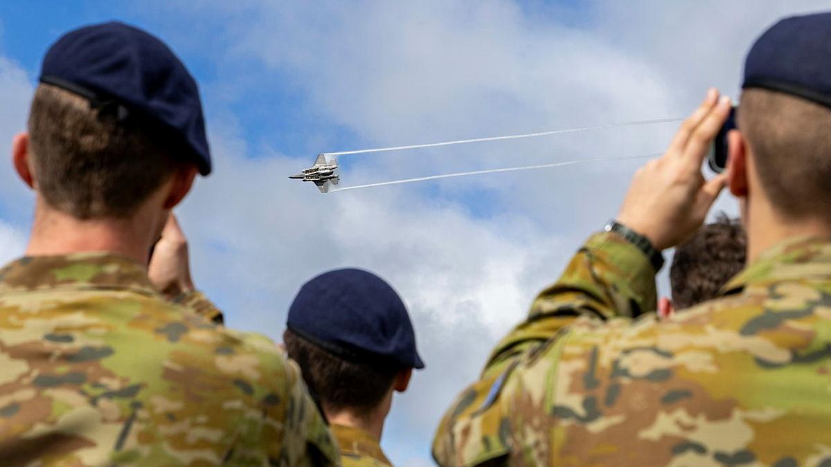 Several military personnel watch an aircraft fly past.