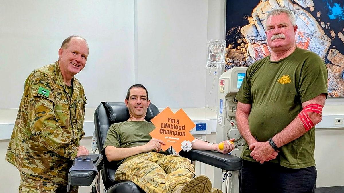 Two soldiers stand either side of another soldier who is donating blood while sitting back in a medical chair.