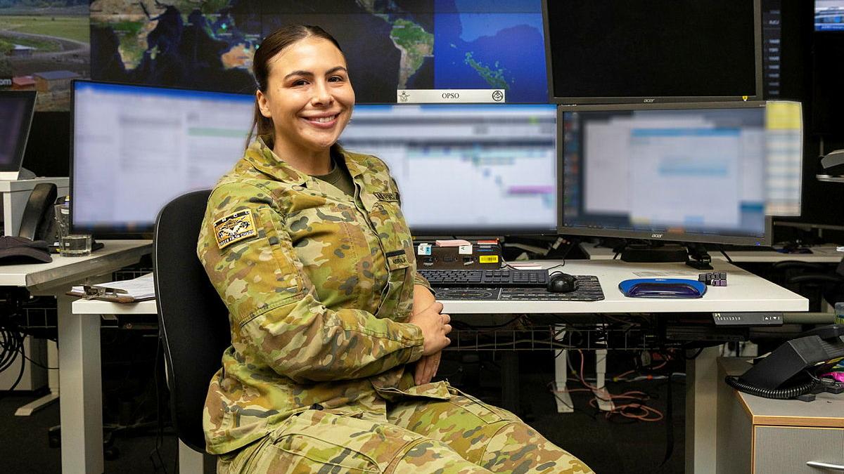 A military woman sitting at a desk with three blurred computer monitors.