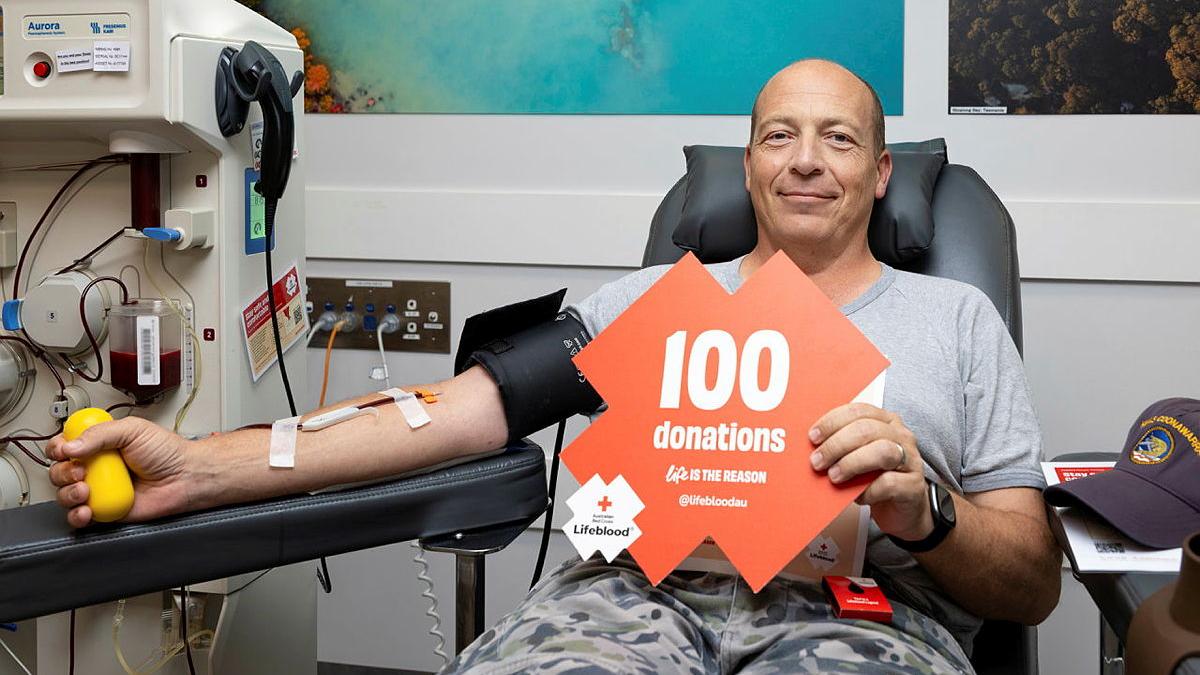 A smiling man sits in a chair and holds a red sign which reads '100 donations'. One of his arms is hooked up to a machine.