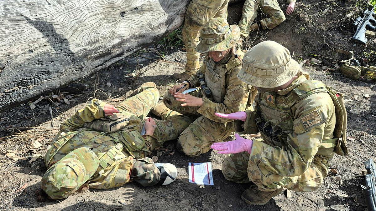 Several military members provide aid to a fallen member in the outdoors.