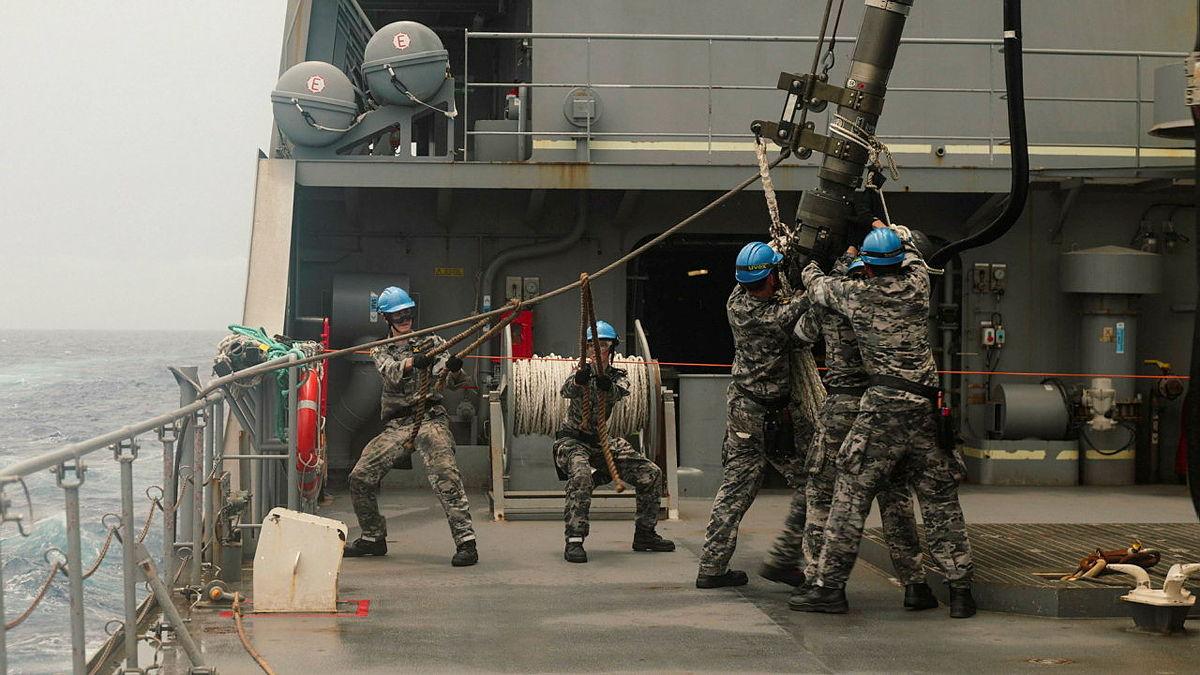 Navy personnel in blue hard hats reach for lines and other equipment on the deck of a ship.