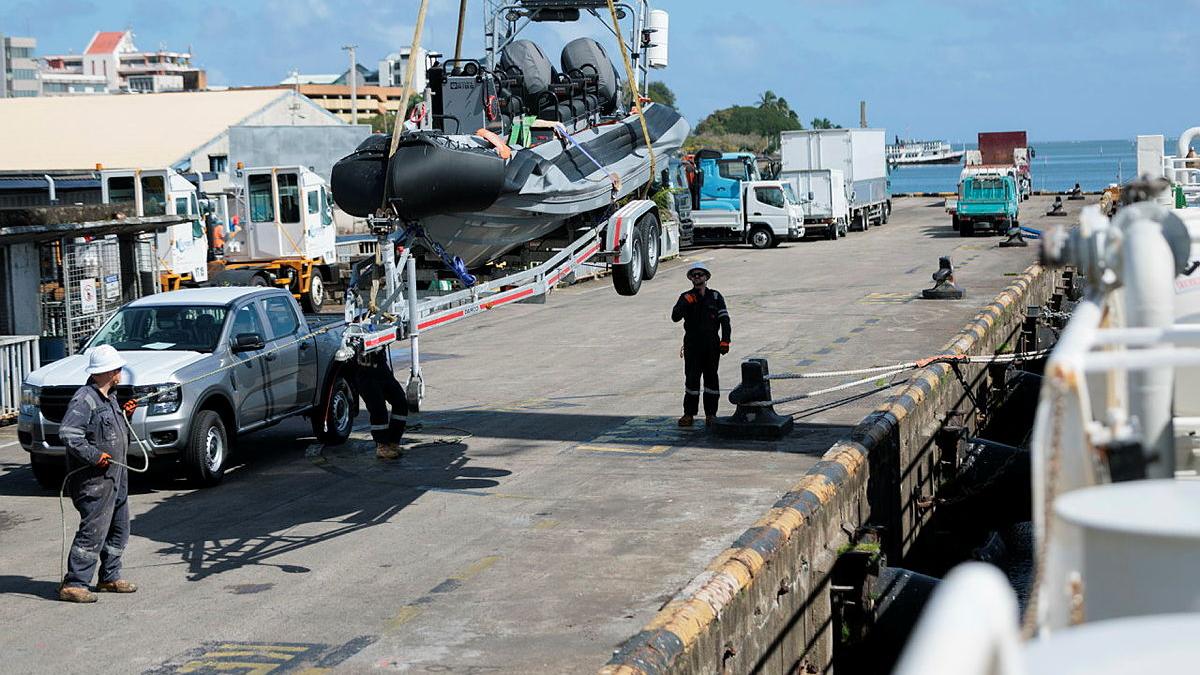 A boat is seen suspended near a ship. People and other vehicles are seen in the vicinity.