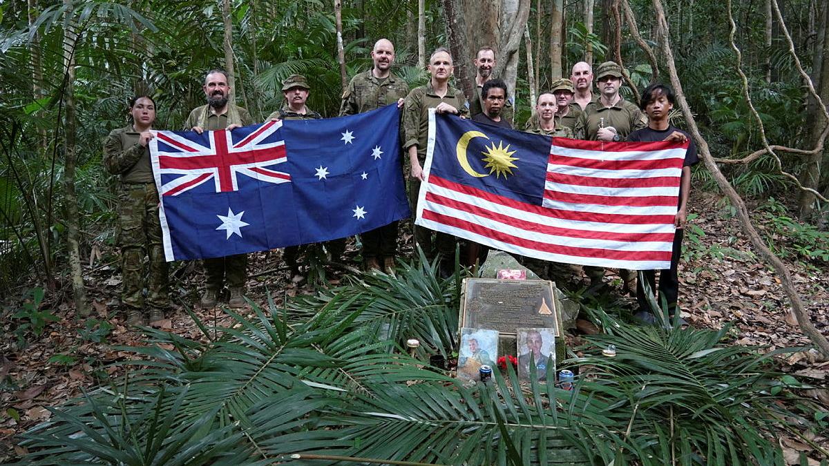 A group of military members hold two flags next to an outdoor tribute decorated with palm leaves.