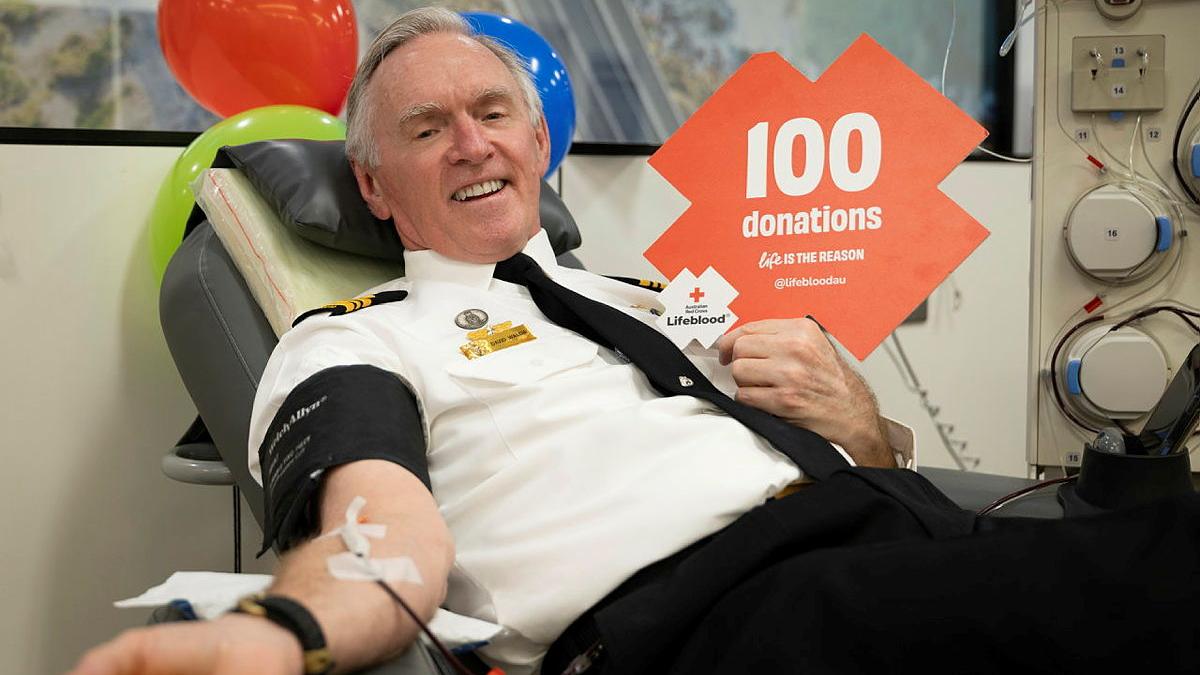 A Navy person in a black and white uniform sits up in a bed with their arm outstretched. They also hold a sign which reads "100 donations".