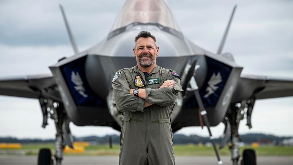 A military man in a flight suit stands before an aircraft on the tarmac.