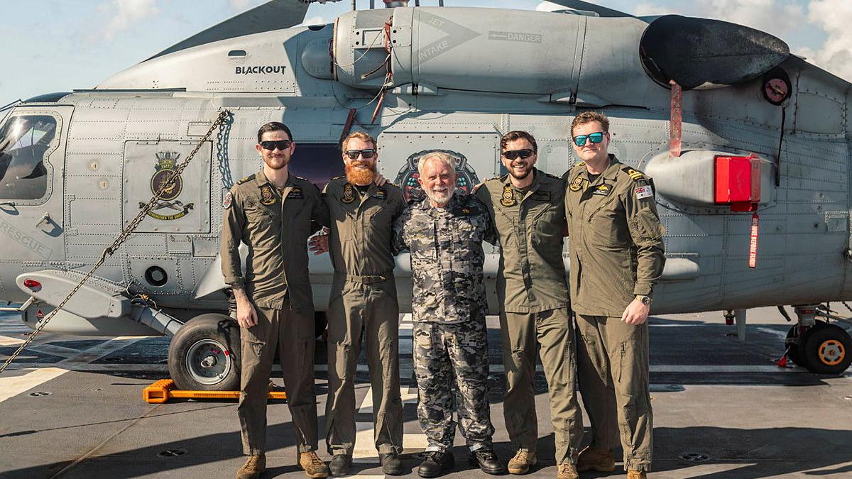 Five people dressed in a mix of flight uniforms and Navy cams stand in front of a helicopter.