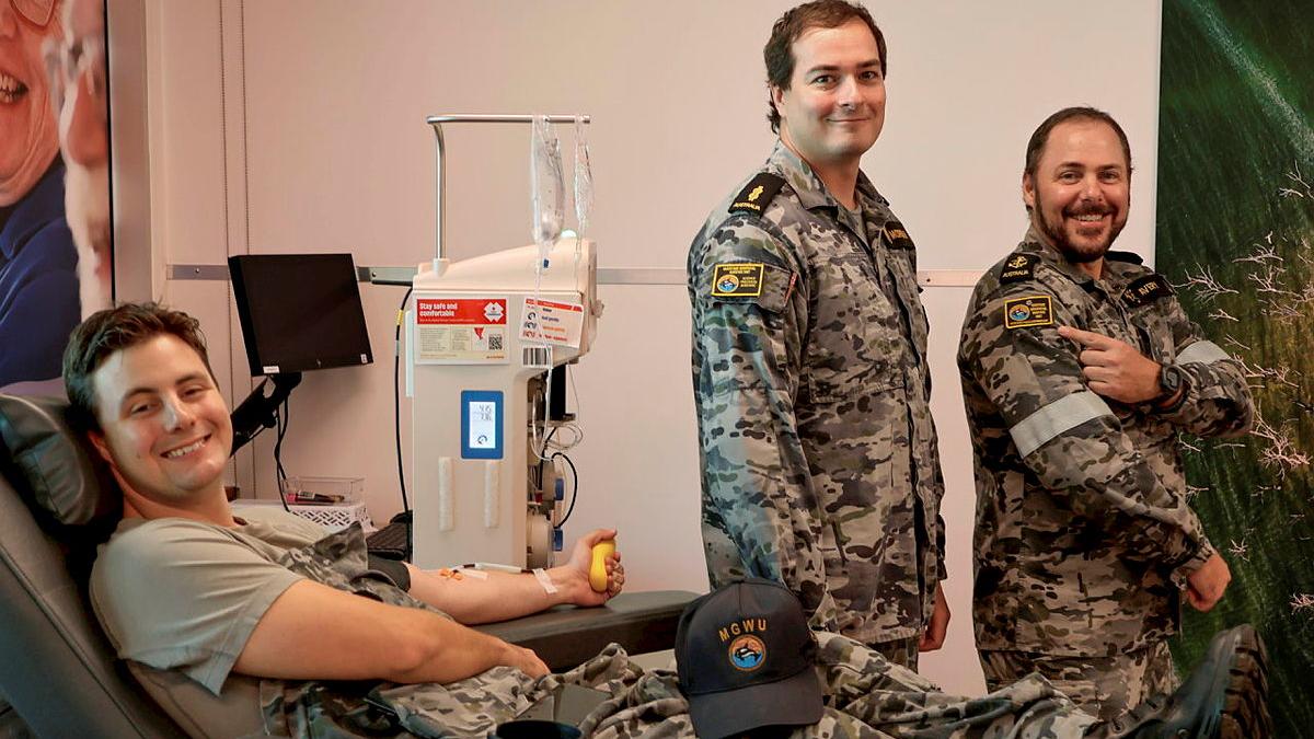 A Navy member sits in a blood donation chair with his arm hooked up to a machine. Two others stand in the room with him.
