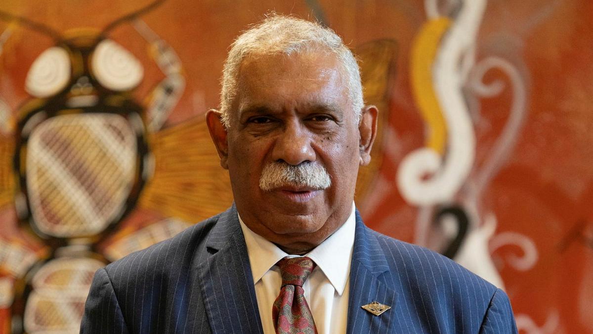 A man in a suit adorned with medals is seen standing in front of an indigenous artwork.