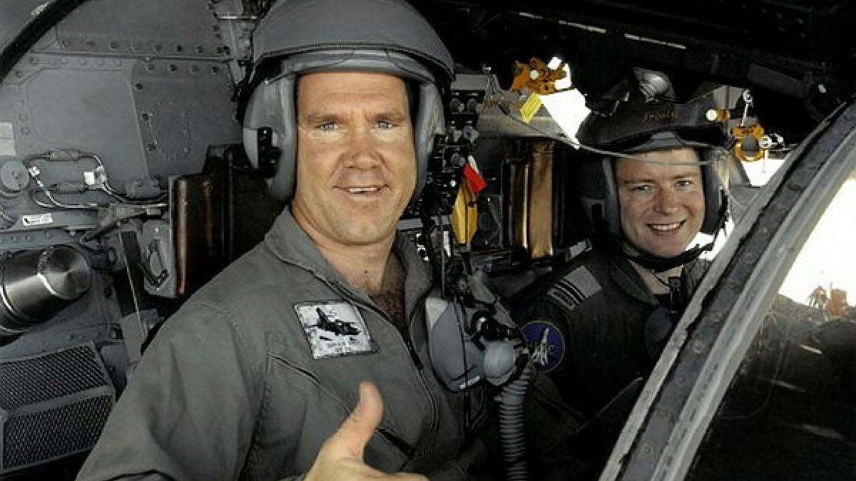 A man in a grey flight suit wears a helmet and gives a 'thumbs up' as he sits in an aircraft next to another person.