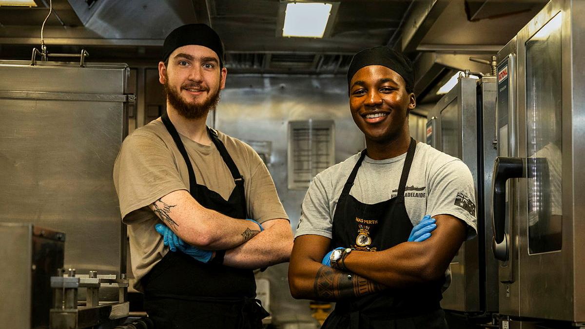Two men in black aprons and headwear stand in a kitchen together with their arms crossed, smiling.