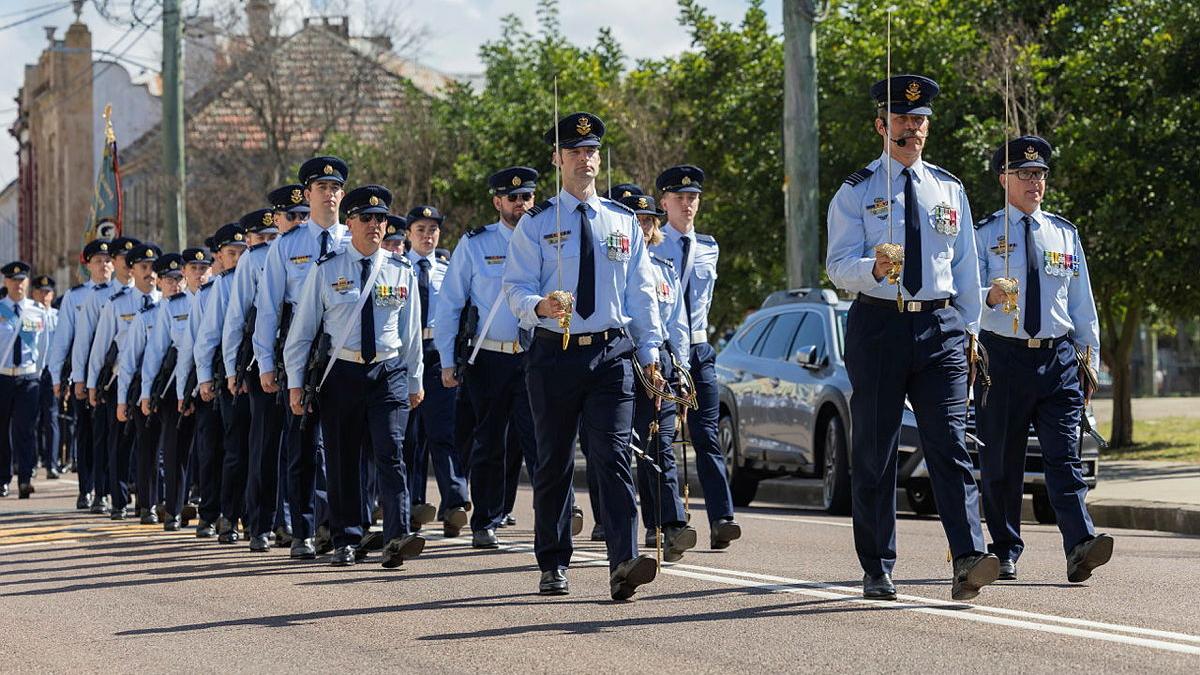 A large group of Air Force personnel with swords held in the air, march together through a city street.