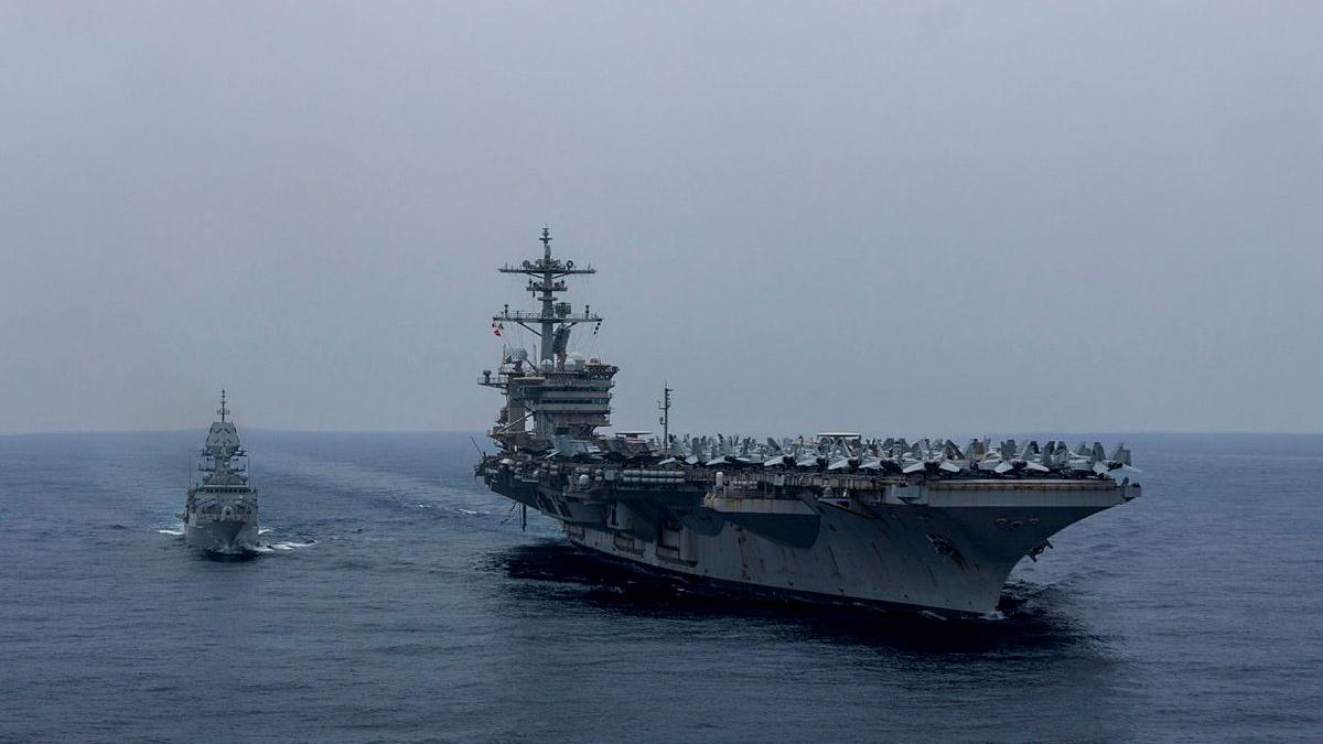 A ship sails close to a much larger vessel which carries numerous aircraft.