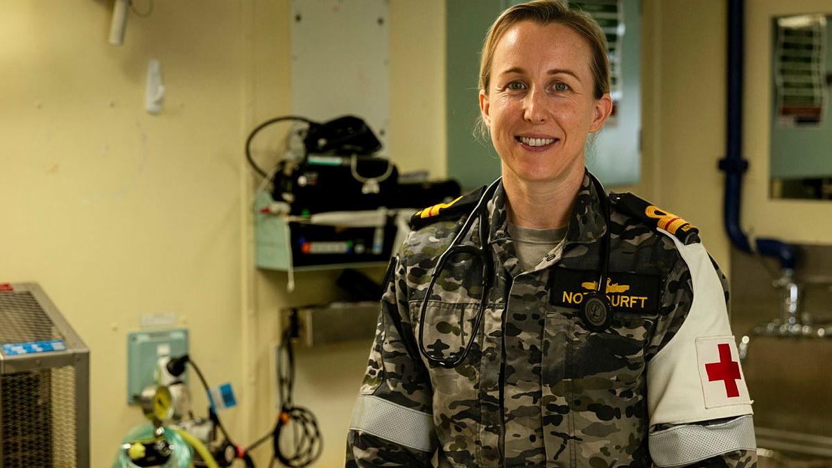 A woman wearing a Navy uniform and a red cross arm band stands in a room of medical equipment.