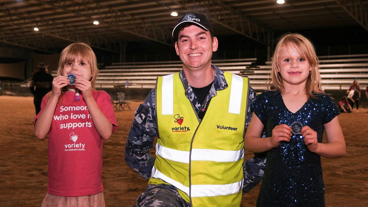 A man in uniform and a high-visibility vest kneels between two young girls in an indoor arena.