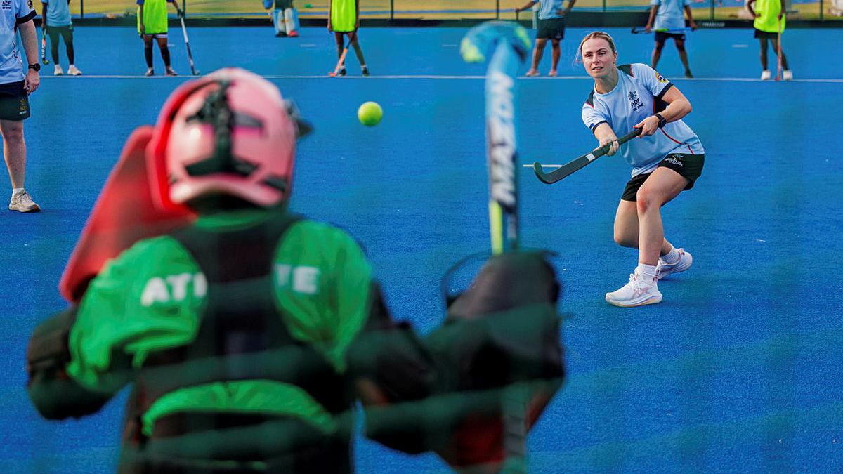 A woman hits a ball with a hockey stick towards a person in protective gear.