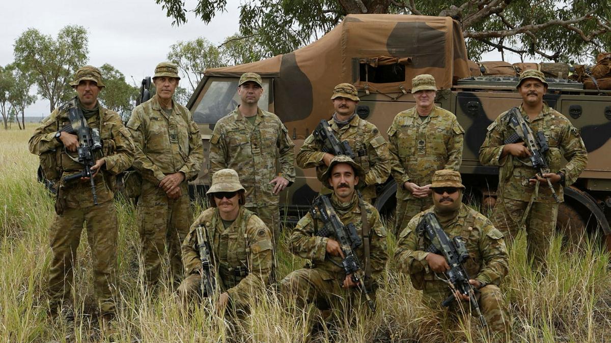 A group of soldiers stand with weapons in front of an Army vehicle in a bush setting.