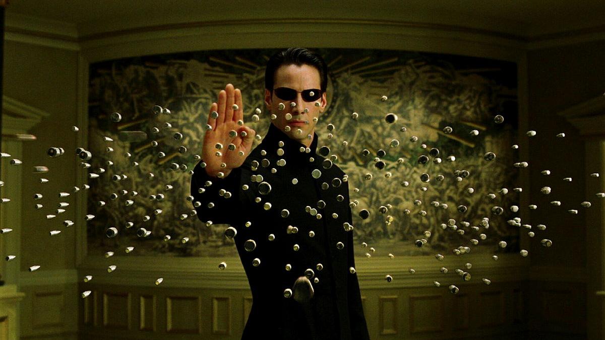 A man wearing sunglasses holds his hand up in front of him as bullets head towards him.